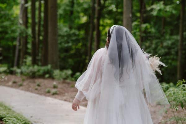 Plus-size bride wearing a flowing white wedding dress with delicate sheer sleeves and a long veil. She is seen from behind, walking along a path in a lush forest setting, exuding an ethereal and romantic aura as she carries a bouquet of dried flowers.