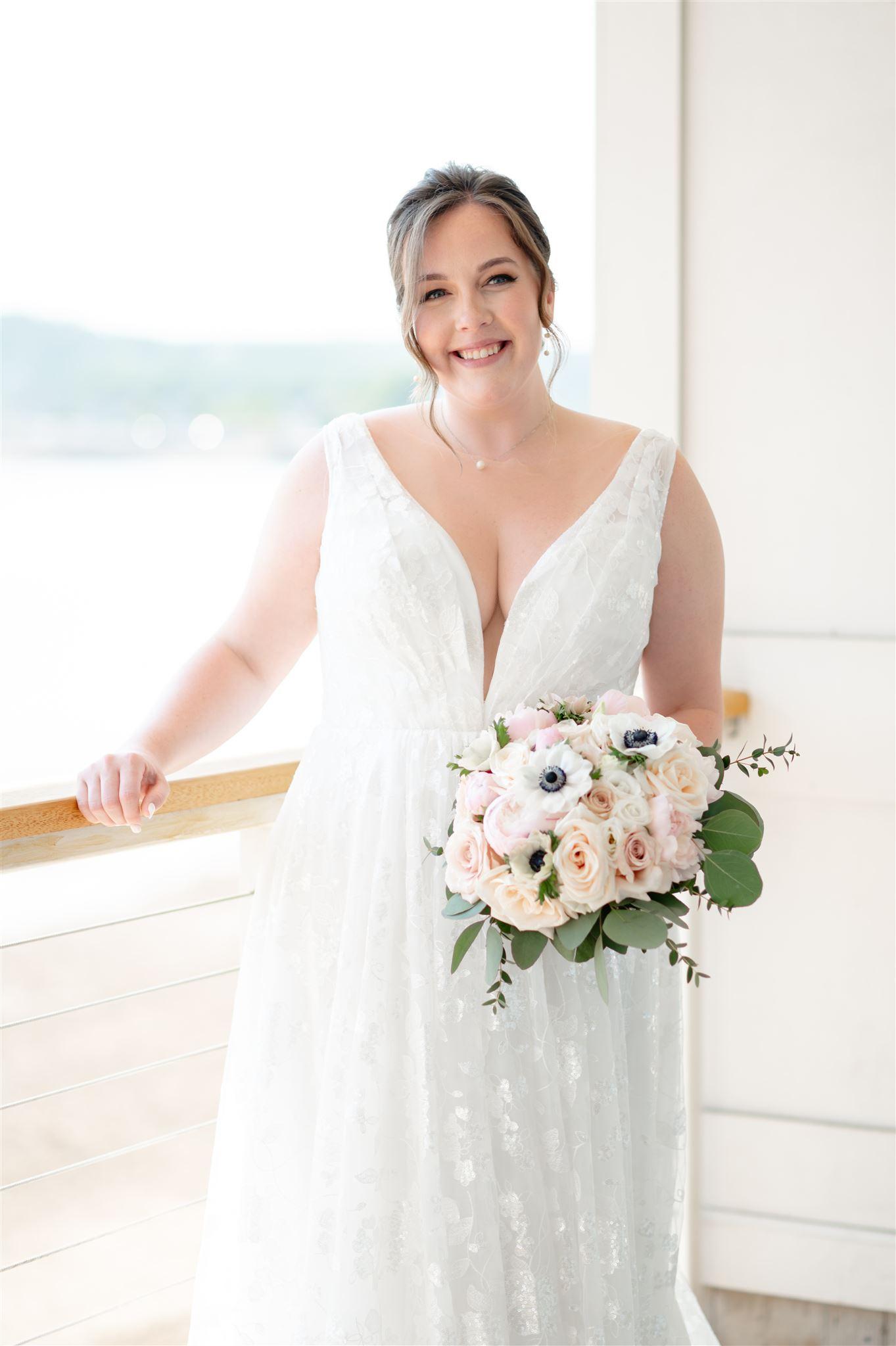 The bride radiates happiness as she poses in her stunning plus-size wedding gown. The deep V-neckline and delicate lace detailing flatter her silhouette, while her bouquet of blush and ivory flowers adds a romantic touch to the serene waterfront setting.