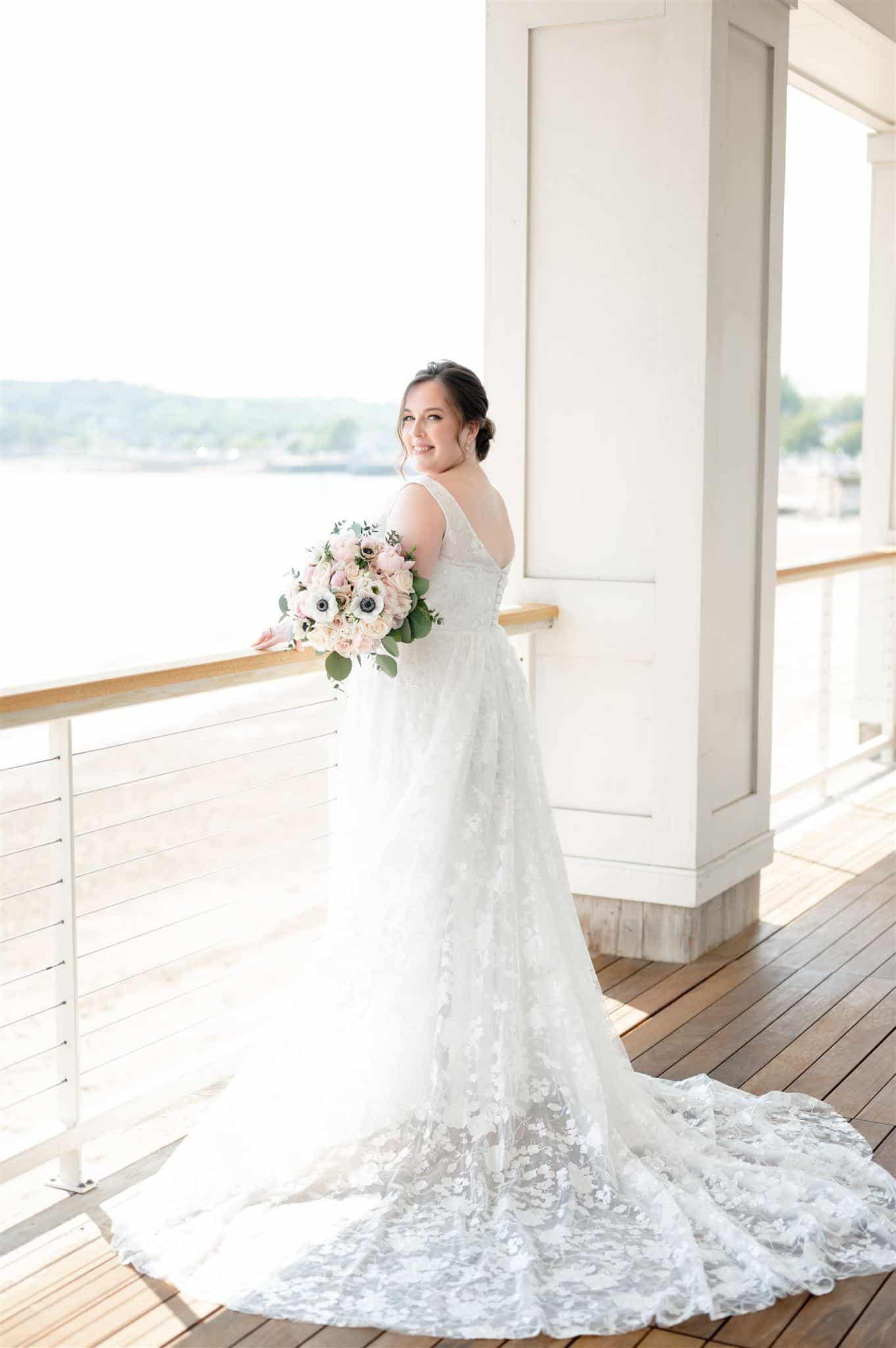 A radiant bride poses gracefully on a seaside balcony, showcasing the intricate lace train of her plus-size wedding gown. The soft floral details and delicate straps beautifully enhance the romantic charm of the dress, perfectly complementing the serene waterfront backdrop.