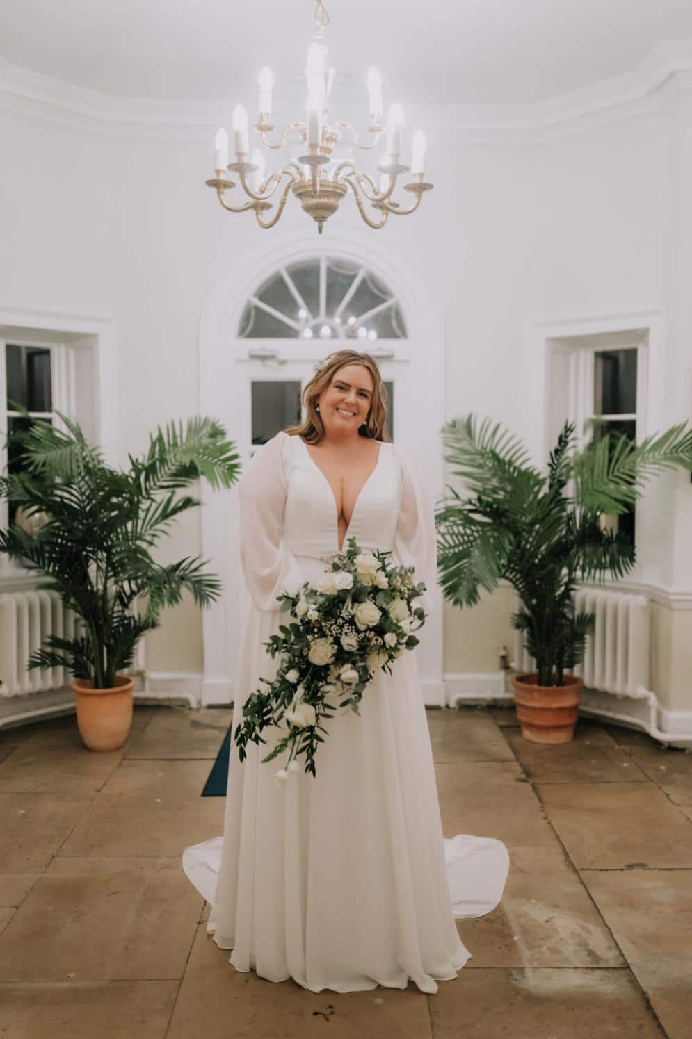 plus-size wedding gown featuring long sleeves, a plunging V-neckline, and a cinched waist with a jeweled belt, perfect for a timeless and sophisticated bridal look