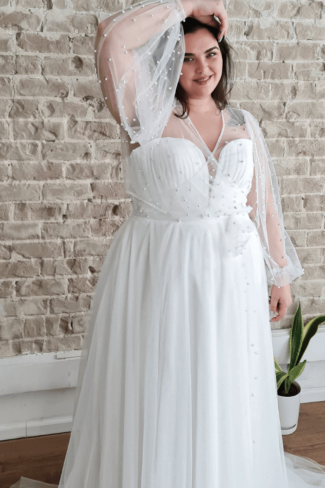 plus-size wedding dress featuring a sweetheart neckline and flowing skirt adorned with delicate pearl details. The gown is complemented by sheer, puffed sleeves with pearl embellishments