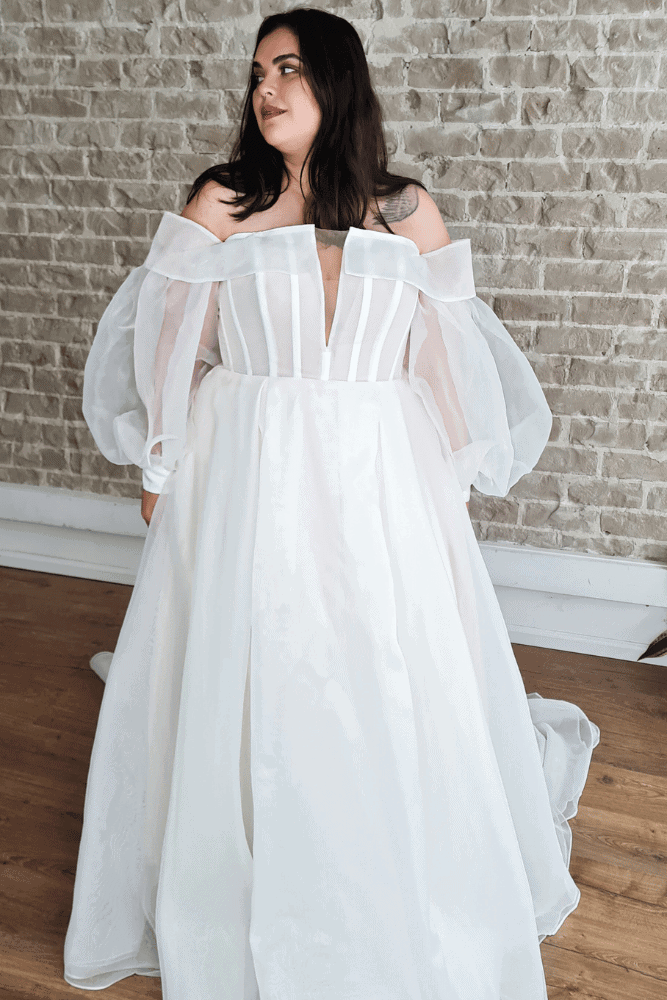 Stylish plus-size wedding dress featuring a fitted bodice with vertical stripes, a deep V-neckline, and dramatic off-the-shoulder sheer sleeves, complemented by a flowing skirt for a modern bridal look.