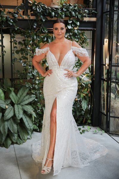 Plus-size model in a sleeveless, beaded plus-size wedding dress with a deep V-neckline, posing in a modern, greenery-filled setting.