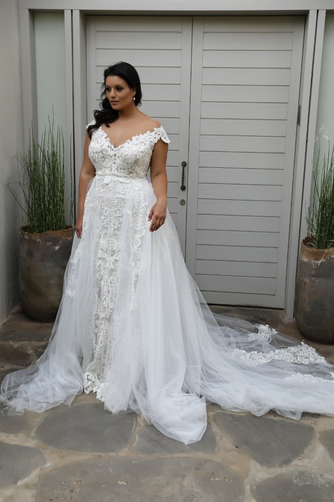 The plus-size bride wears a lace wedding gown with intricate detailing on the back and a long tulle train that cascades onto the ground