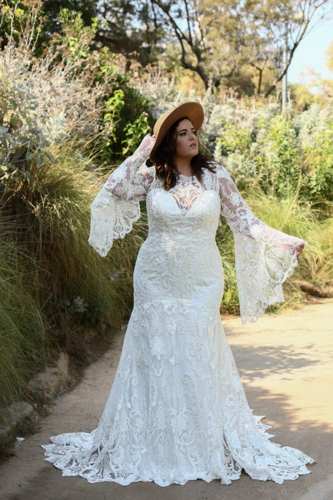 Plus-size bride in a lace wedding dress with long bell sleeves and a detailed train, wearing a wide-brimmed hat and standing on a scenic pathway surrounded by lush greenery.