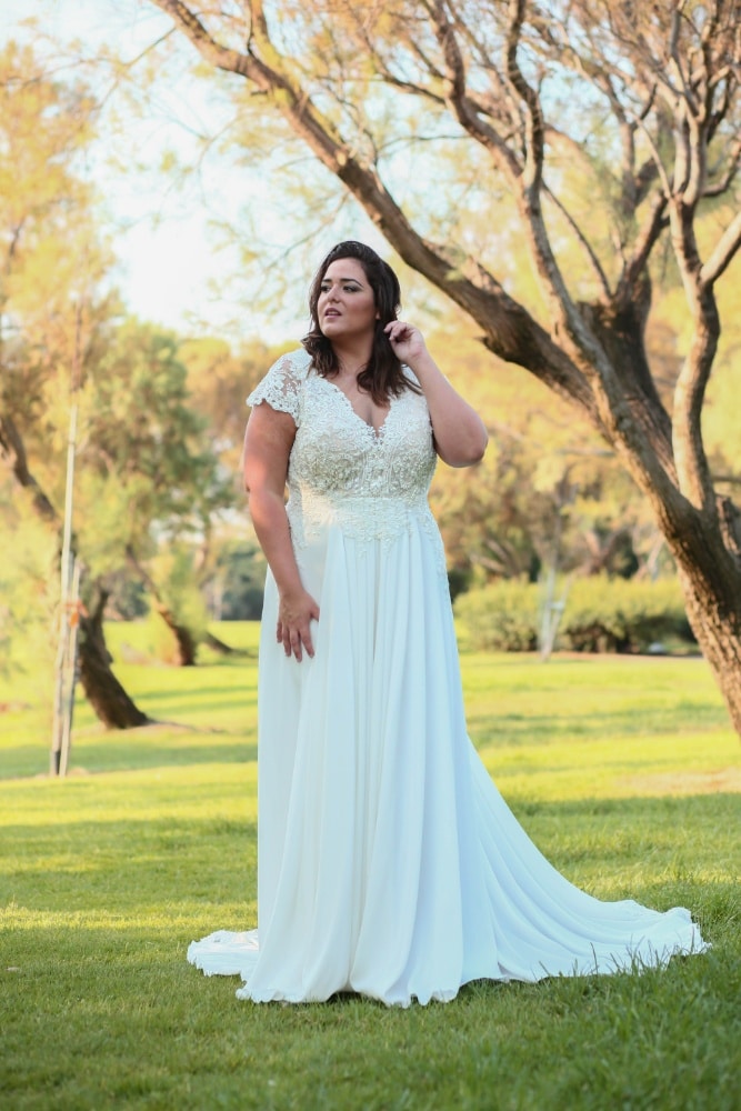 Plus-size bride wearing a flowing wedding dress with a lace bodice and cap sleeves, standing gracefully on a lush green lawn with trees in the background.