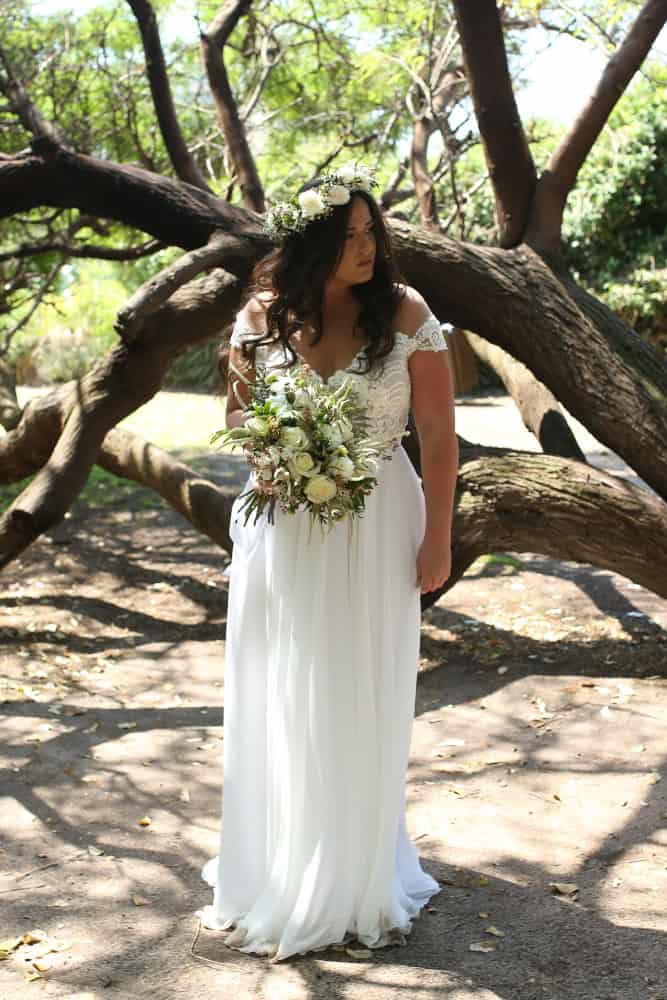 Plus-size bride in a lace wedding dress with off-the-shoulder sleeves, wearing a floral crown and holding a lush bouquet, standing beneath the branches of a large, twisted tree.