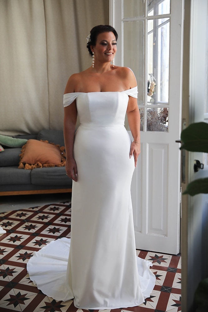 Radiant plus-size bride exuding confidence in a sleek, minimalist off-the-shoulder wedding gown. The fitted silhouette and clean lines beautifully enhance her figure