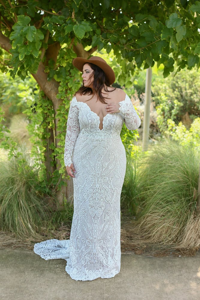 Plus-size bride in a detailed lace wedding dress with an off-the-shoulder neckline and a flowing train, standing on a sunlit path surrounded by lush greenery.