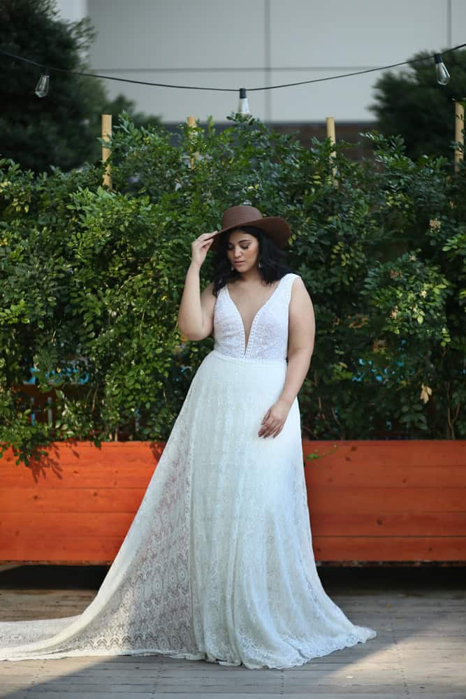 Plus-size bride wearing a lace wedding dress with a plunging neckline and a cowboy hat, standing confidently against a backdrop of greenery and string lights.
