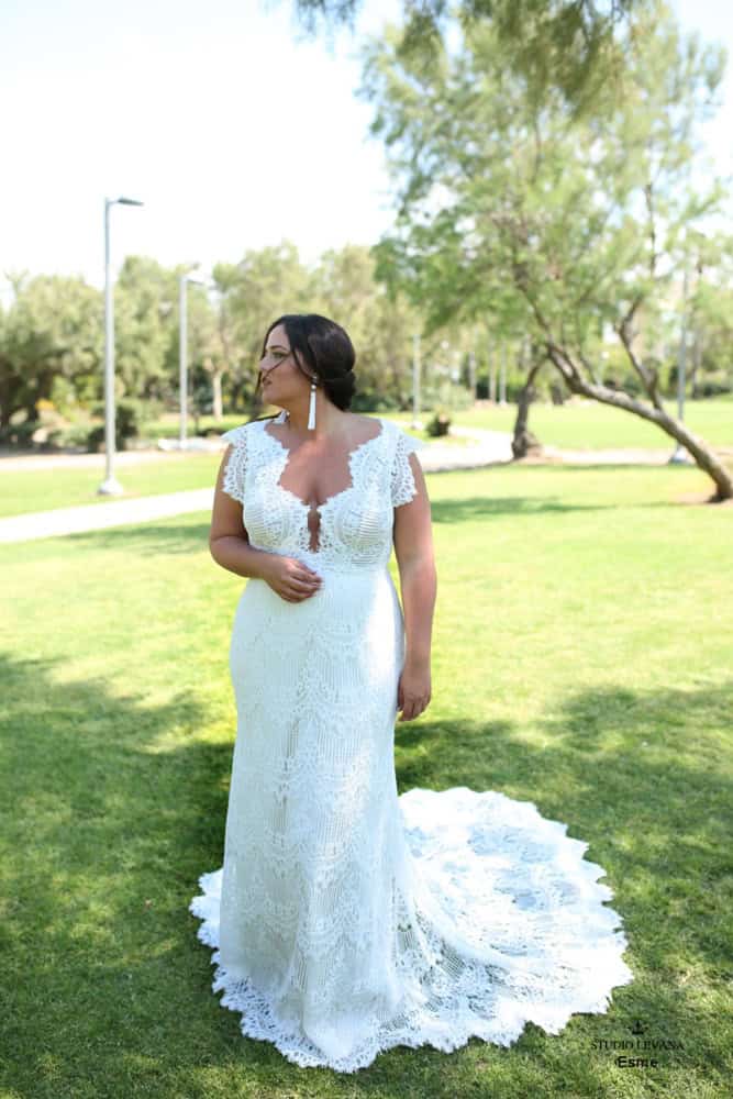 Plus-size bride wearing a form-fitting lace wedding dress with a scalloped train and deep neckline, standing in a sunlit park surrounded by greenery.