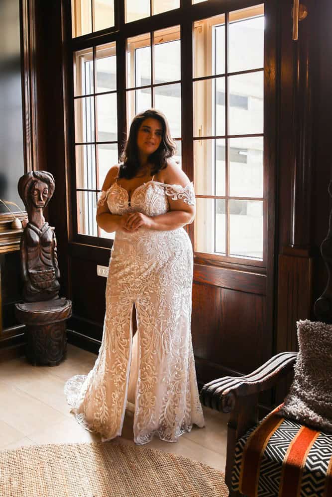 Plus-size bride in a stunning lace gown with off-the-shoulder straps and a fitted silhouette, standing gracefully near a window with warm, natural light streaming in.
