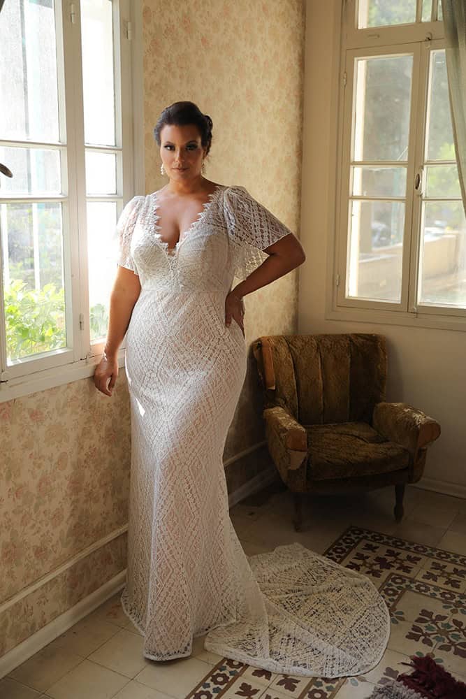Elegant plus-size bride in a beautiful lace wedding gown featuring flutter sleeves and a deep V-neckline. The geometric lace design and form-fitting silhouette beautifully highlight her figure