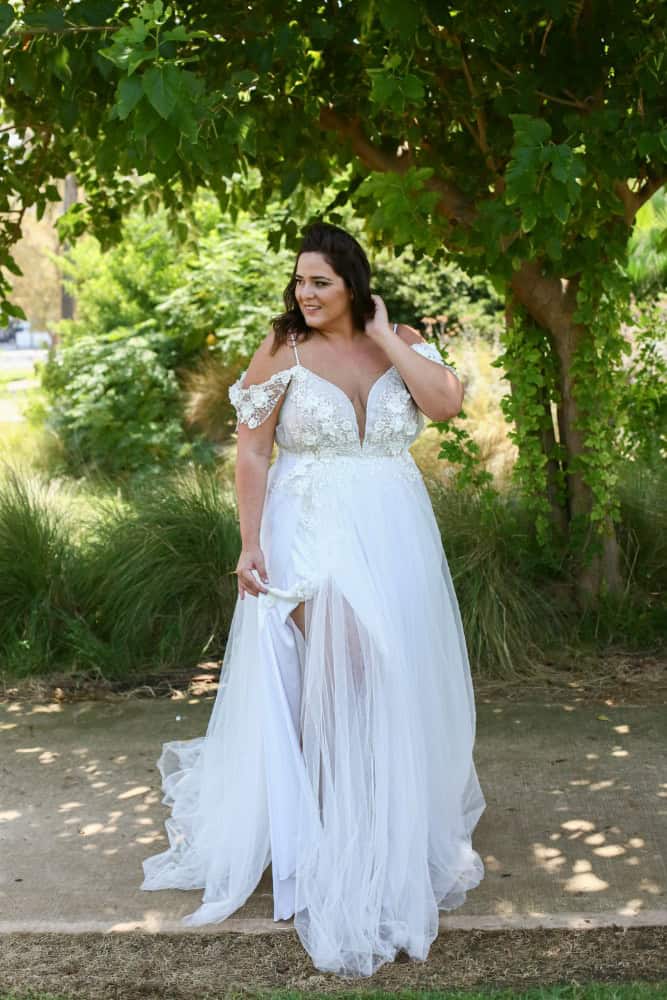 Plus-size bride in a flowing wedding dress with lace details and cap sleeves, standing gracefully under the branches of a large tree in a sunlit woodland setting.