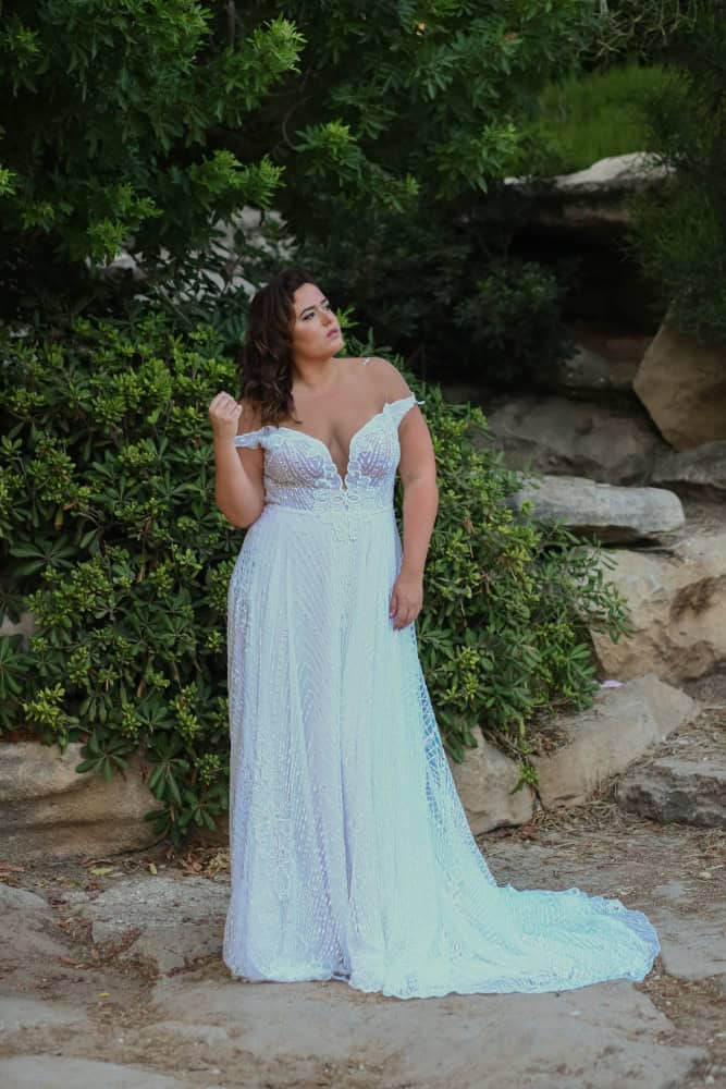 Plus-size bride wearing an off-the-shoulder lace wedding dress with a plunging neckline, standing elegantly in front of lush green foliage and natural rock formations.
