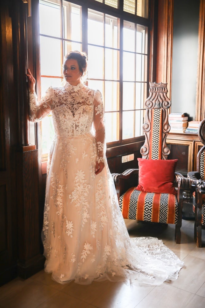 The plus-size bride, adorned in a lace wedding gown with sheer long sleeves, a high neckline, and floral embroidery, stands gracefully by a window.