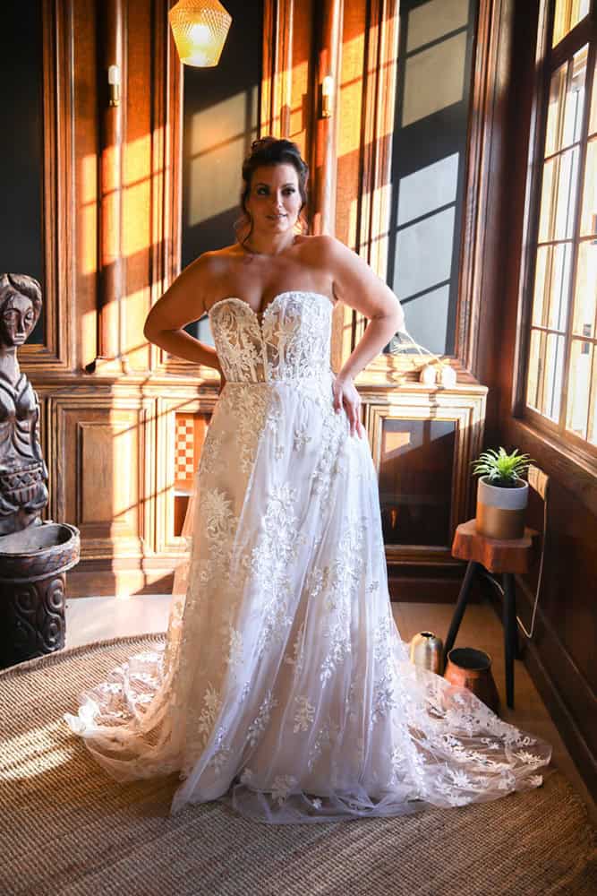 This strapless plus-size gown showcases a structured, corset-inspired bodice adorned with intricate lace appliques. The flowing skirt cascades elegantly, accented by delicate floral patterns, offering a romantic and regal bridal look.