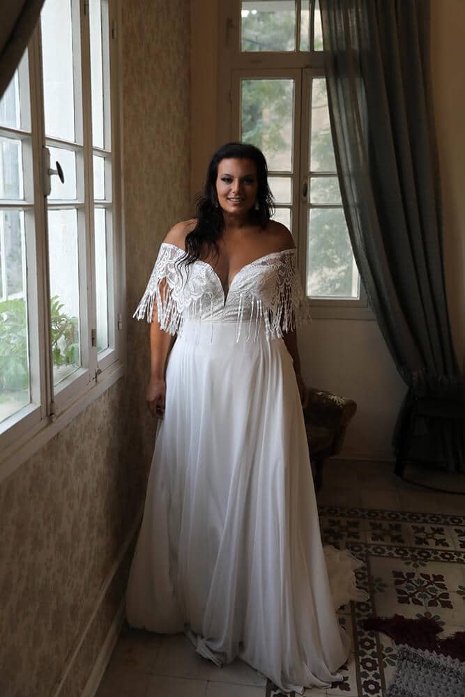 Stunning plus-size bride in an off-the-shoulder wedding gown adorned with intricate lace and playful fringe details. The flowing chiffon skirt adds a touch of elegance