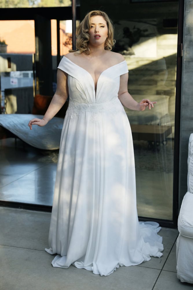 Plus-size bride in an off-the-shoulder wedding dress with a deep plunging neckline and delicate lace detailing, standing in a softly lit, wooded setting.