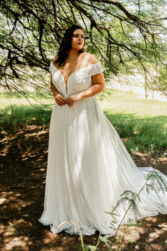 Plus-size bride in a sparkling off-the-shoulder wedding gown with a deep neckline, standing gracefully under a canopy of lush tree branches.