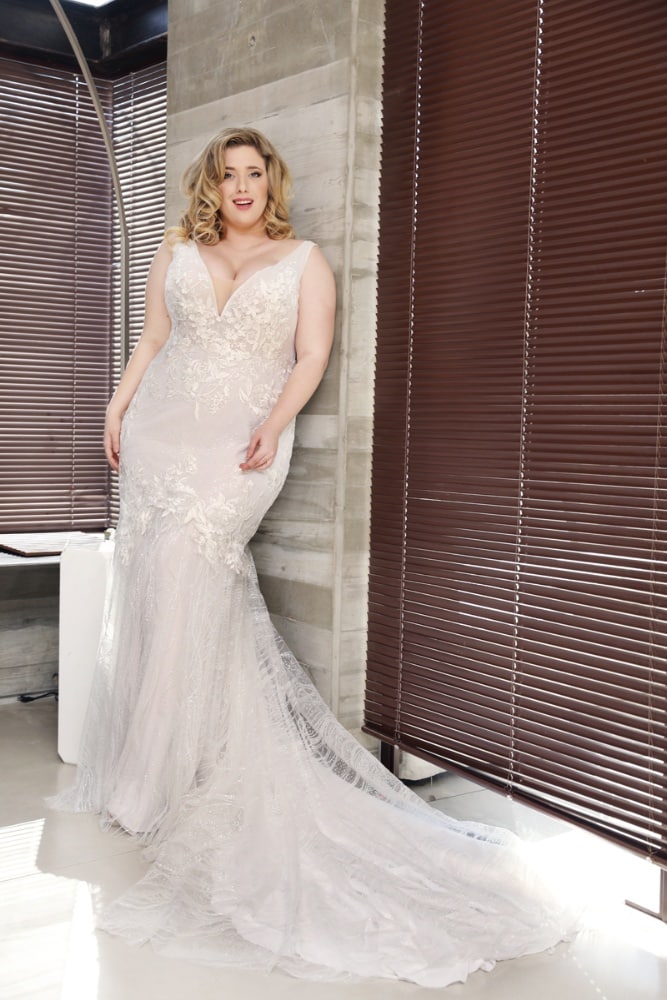 Plus-size bride wearing a fitted lace mermaid wedding dress with a long train, posing elegantly against a modern concrete wall with blinds filtering the light.
