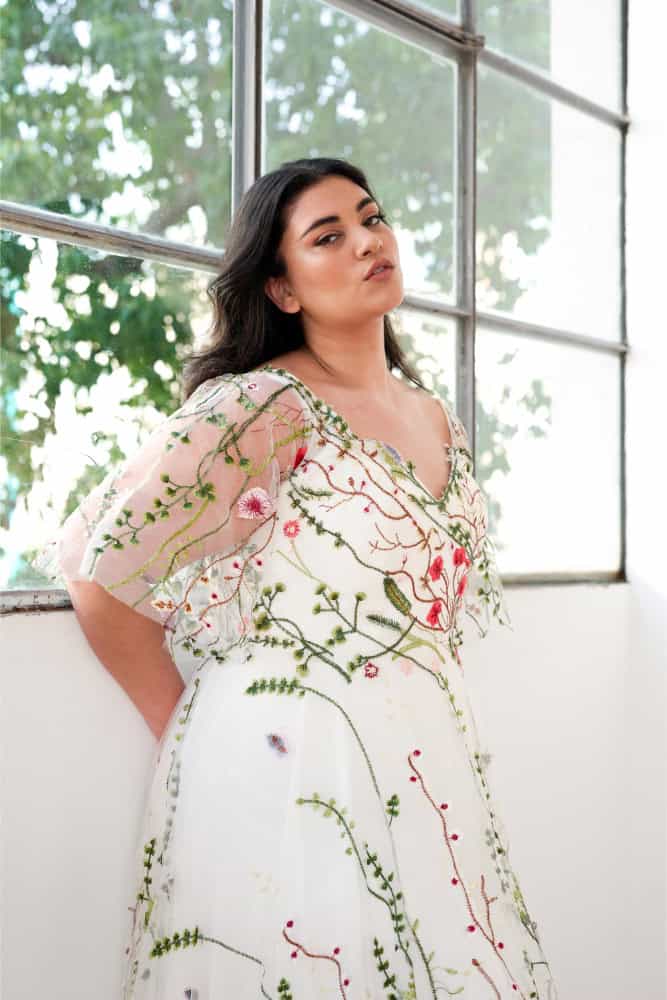 Plus-size bride wearing a stunning plus-size wedding dress with an invisible internal corset, featuring a flowing silhouette adorned with intricate floral embroidery and delicate sheer sleeves. The gown showcases a flattering V-neckline