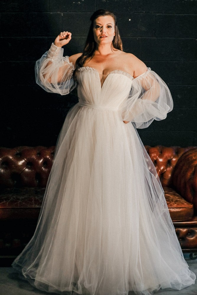 A plus-size bride wearing a strapless, off-the-shoulder tulle wedding gown with dramatic puff sleeves