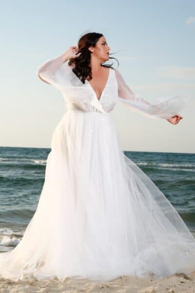 Plus-size bride wearing a sparkling, long-sleeved white gown, standing on the sandy beach with the sea in the background