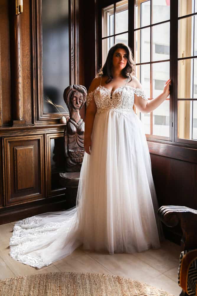 Plus-size bride in a romantic off-the-shoulder wedding gown with intricate lace detailing and a flowing tulle skirt, standing by a large window.