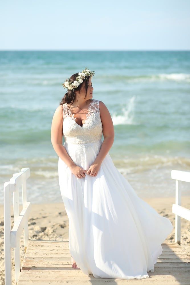 "Plus-size bride facing the ocean, wearing a flowing white gown with lace details and a floral crown, standing on a wooden pathway leading to the beach."