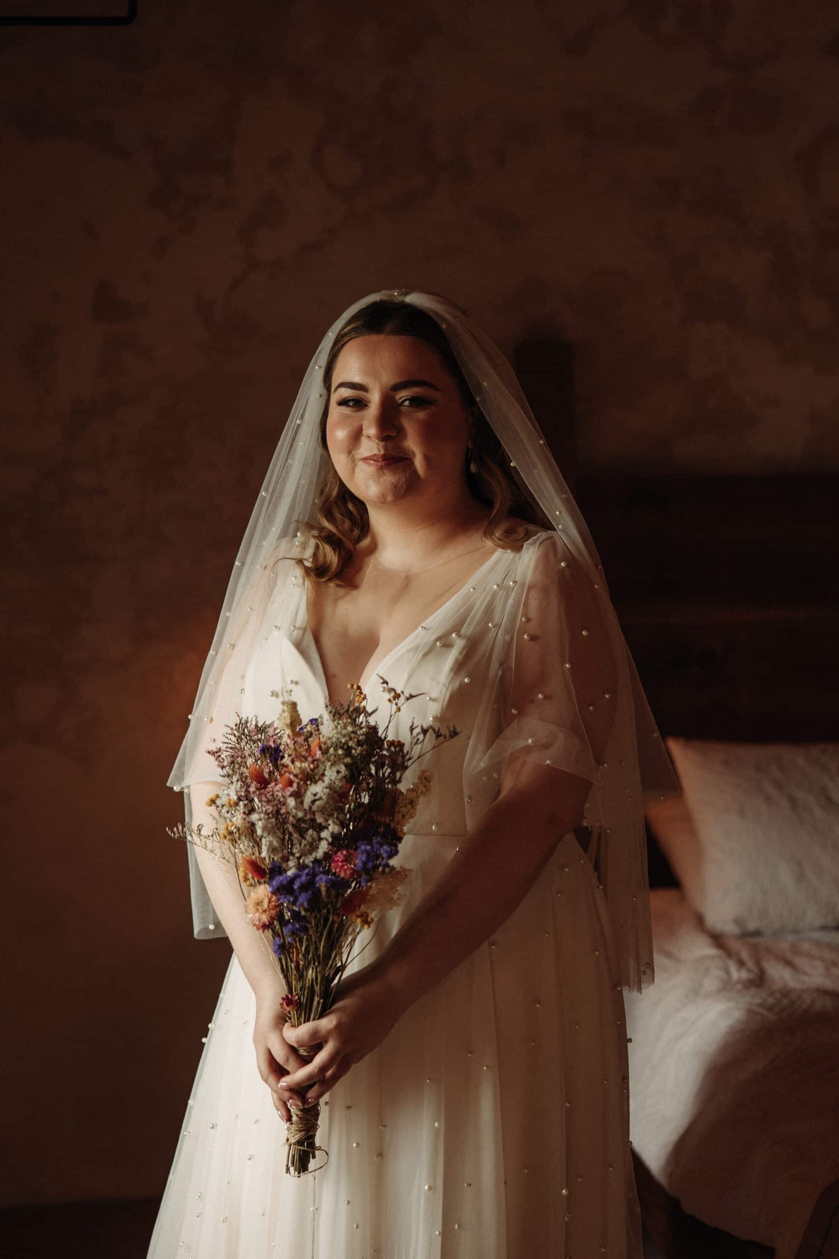The bride, in a pearl-embellished dress and veil, holds a bouquet of dried flowers, standing in a dimly lit room.