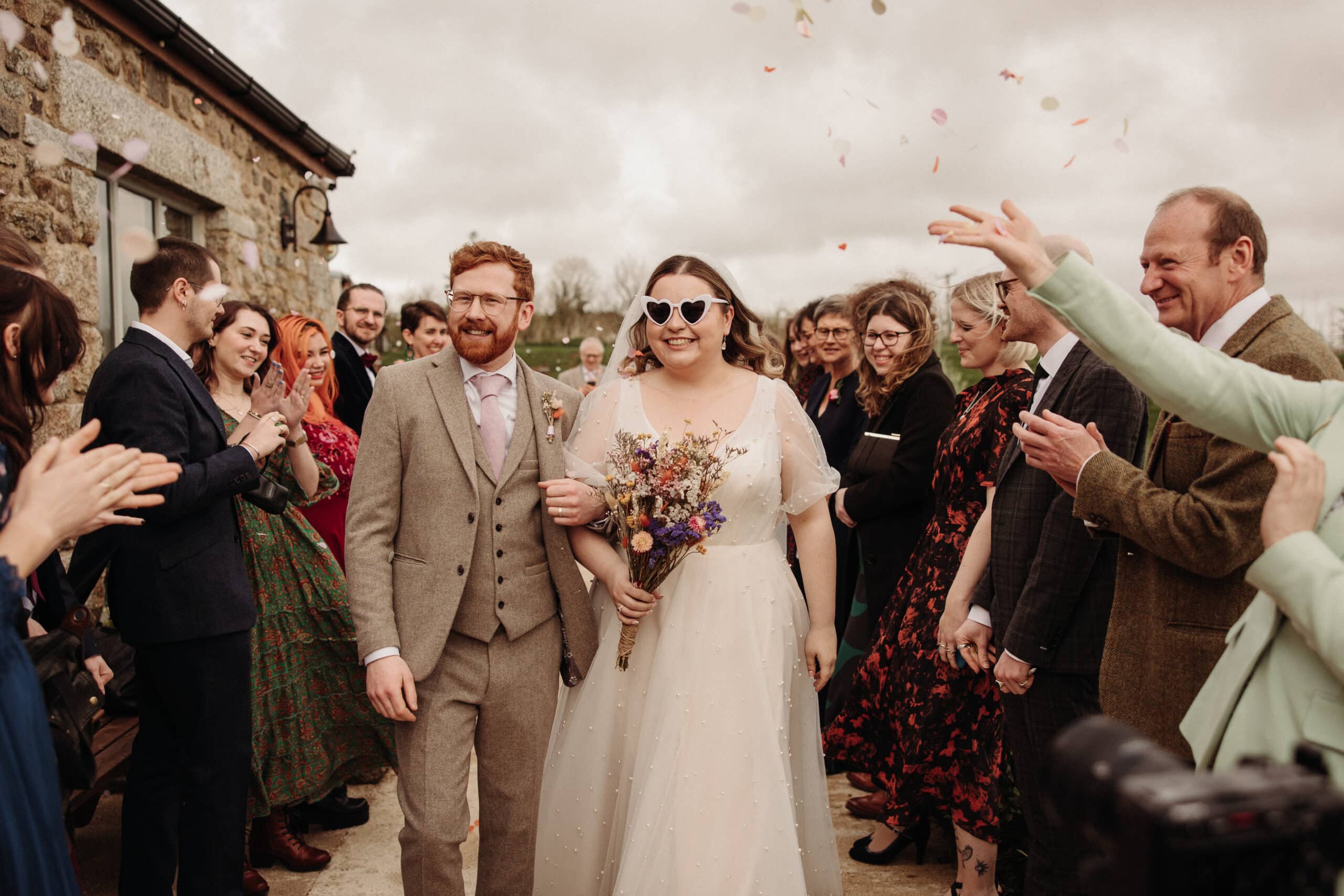A bride in a white dress and sunglasses poses with the groom in a tweed suit against a wooden backdrop.