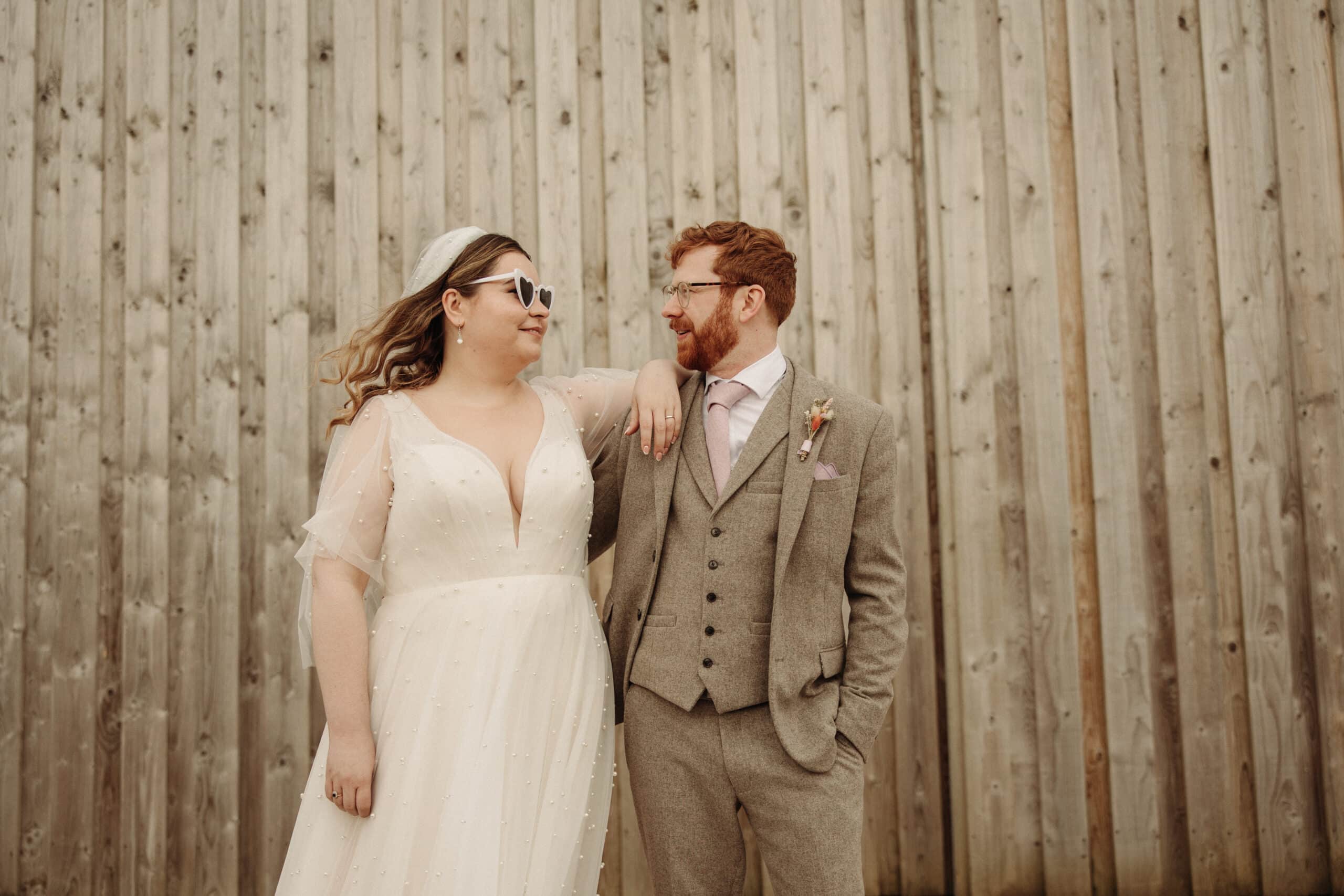 A bride in a white dress and sunglasses poses with the groom in a tweed suit against a wooden backdrop.