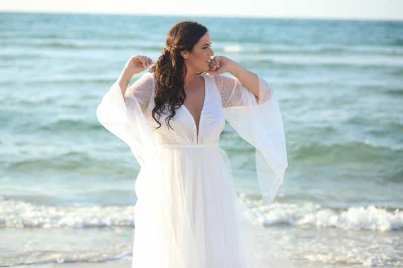 The plus-size bride, wearing a white boho gown with sheer, fluttering sleeves and a deep V-neck, stands gracefully on the beach.