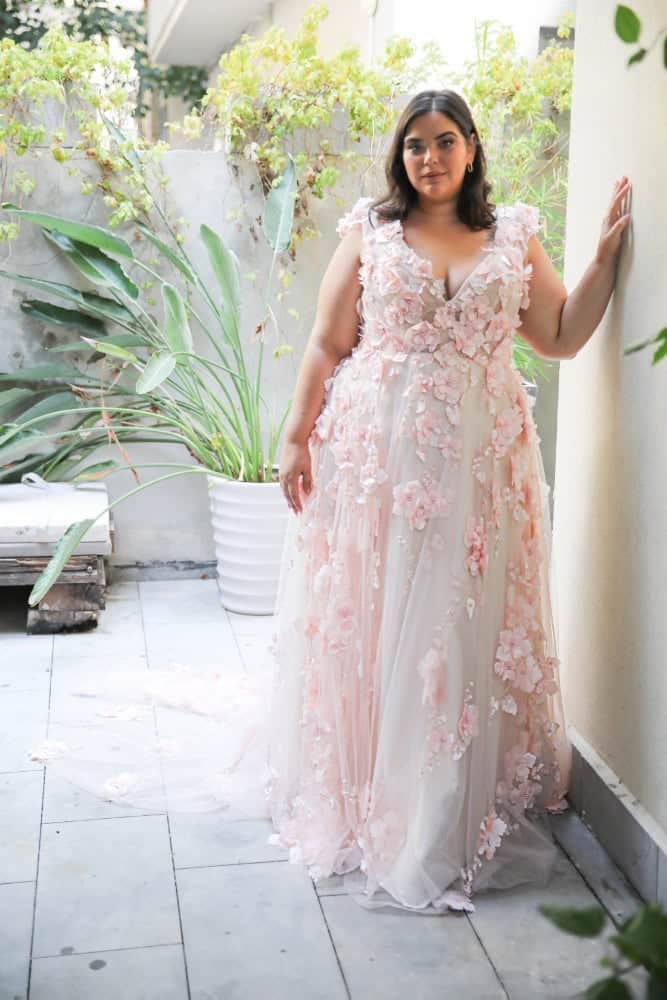 Plus-size bride in a blush pink wedding gown adorned with 3D floral appliqués and a flowing train,