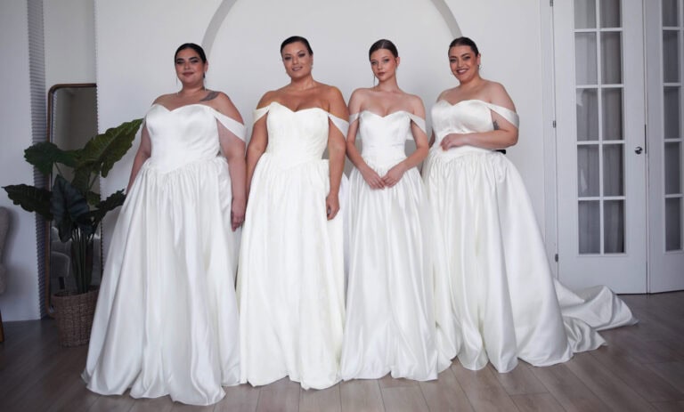 Four women of various body types, dressed in elegant white wedding gowns, stand side by side in a modern, well-lit room with minimalist decor. Each gown features unique design elements like off-shoulder necklines and fitted bodices, emphasizing a contemporary bridal style suitable for all sizes.