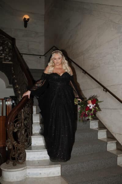 "Plus-size bride showcases a stunning customized black wedding gown with intricate lace detailing and dramatic off-the-shoulder sleeves, descending an elegant staircase."