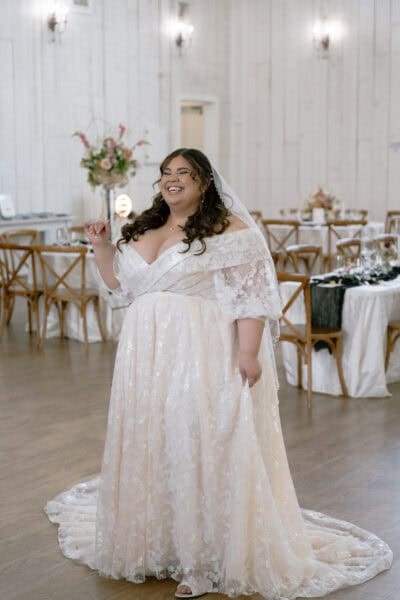 Radiant bride wearing a custom plus-size wedding dress with off-the-shoulder floral lace details, sparkling fabric, and a flowing train, celebrating in a beautifully decorated venue.