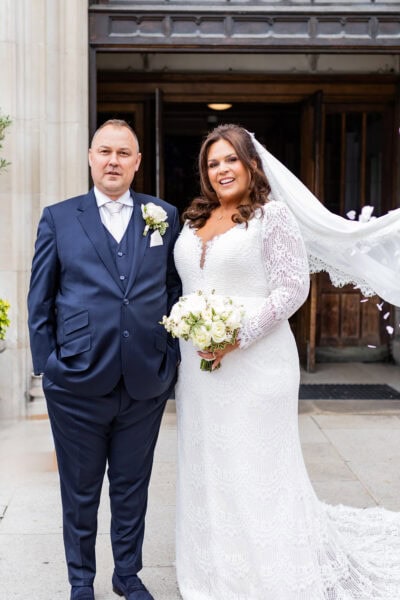 Bride in a stunning lace wedding dress with long sleeves and a plunging neckline, standing beside her groom, holding a bouquet of white flowers.