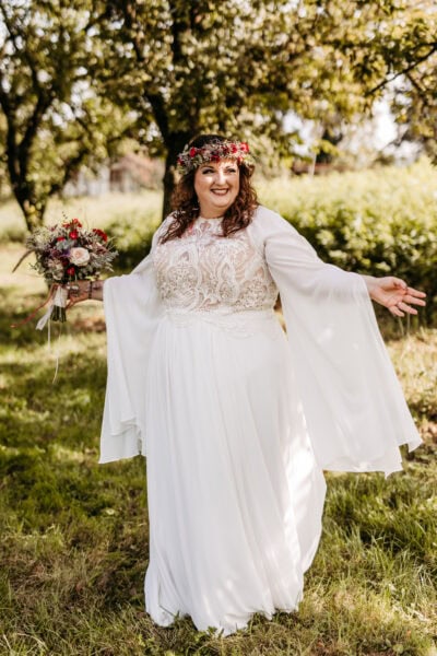 Jutta in the Emma gown by Studio Levana, a plus-size boho wedding dress with cape sleeves and a flower wreath, radiating boho elegance in her countryside wedding.