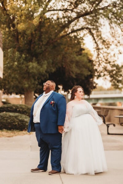 A joyful couple posing outdoors, the bride glowing in her custom plus size bridal gown and the groom in a sharp blue suit.