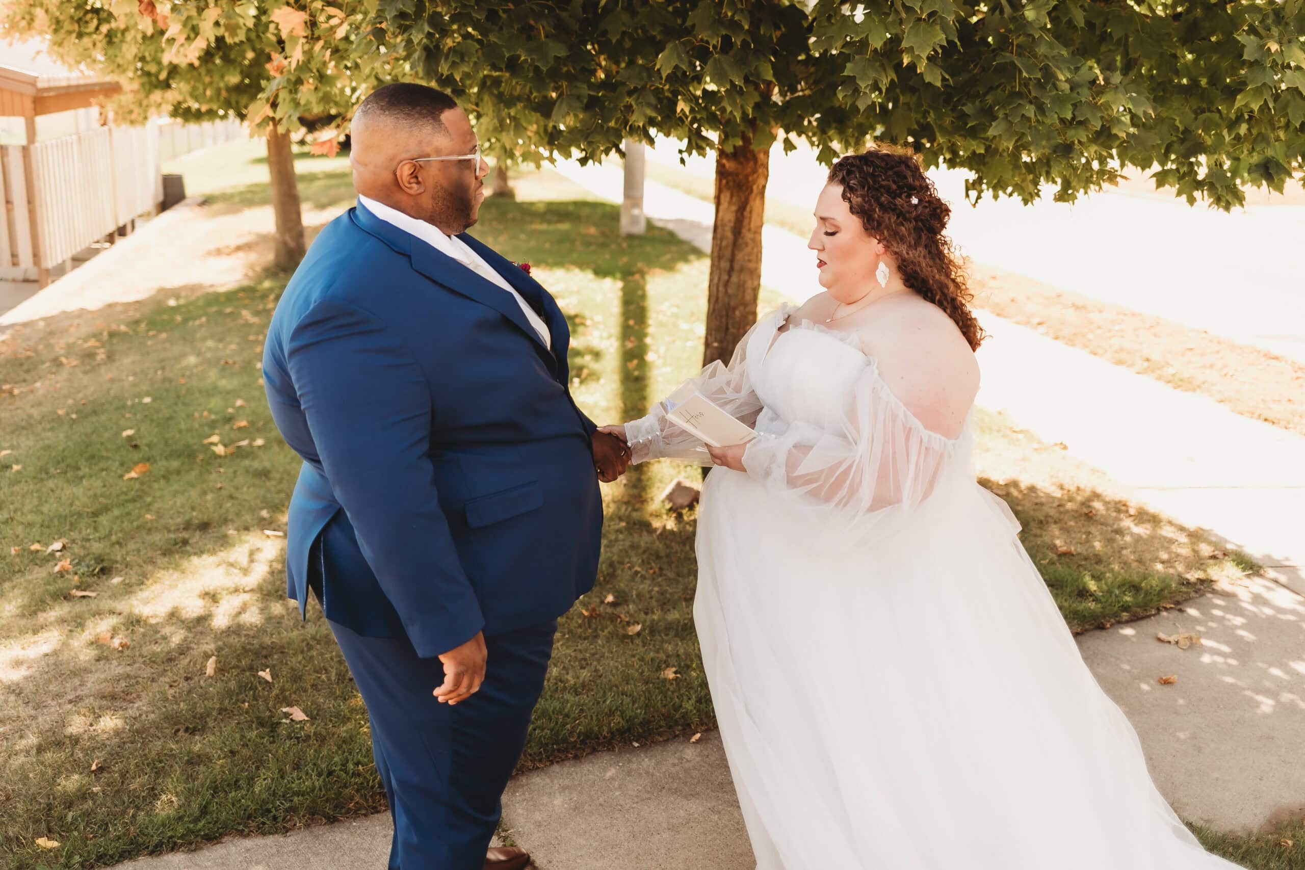 A sweet moment of the couple exchanging personal vows, with the bride radiant in her plus size bridal gown under a leafy backdrop.
