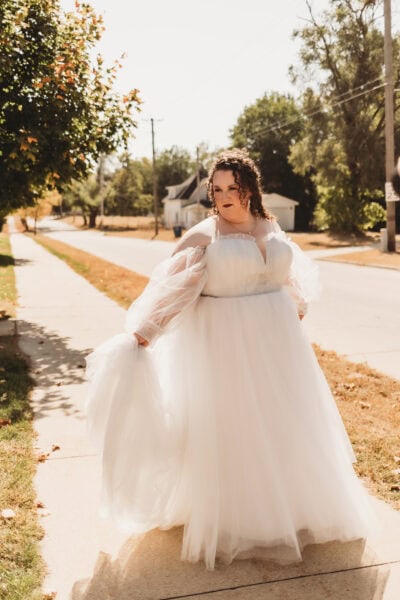 A radiant bride wearing a flowing plus size bridal gown, showcasing elegant off-the-shoulder tulle sleeves and a confident smile.