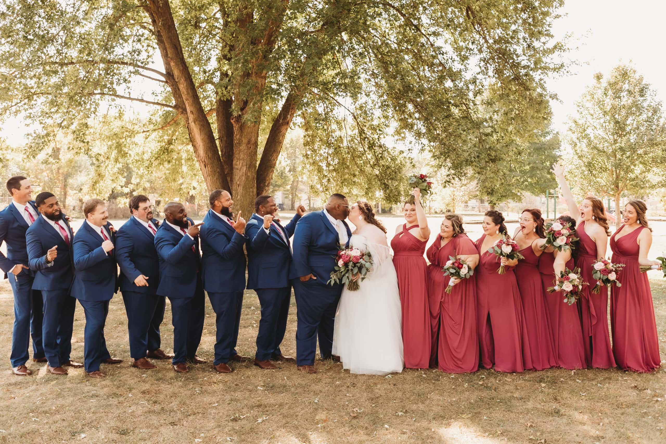 A joyous wedding party surrounds the bride in her elegant plus size bridal gown and the groom in a blue suit as they share a kiss under a tree.