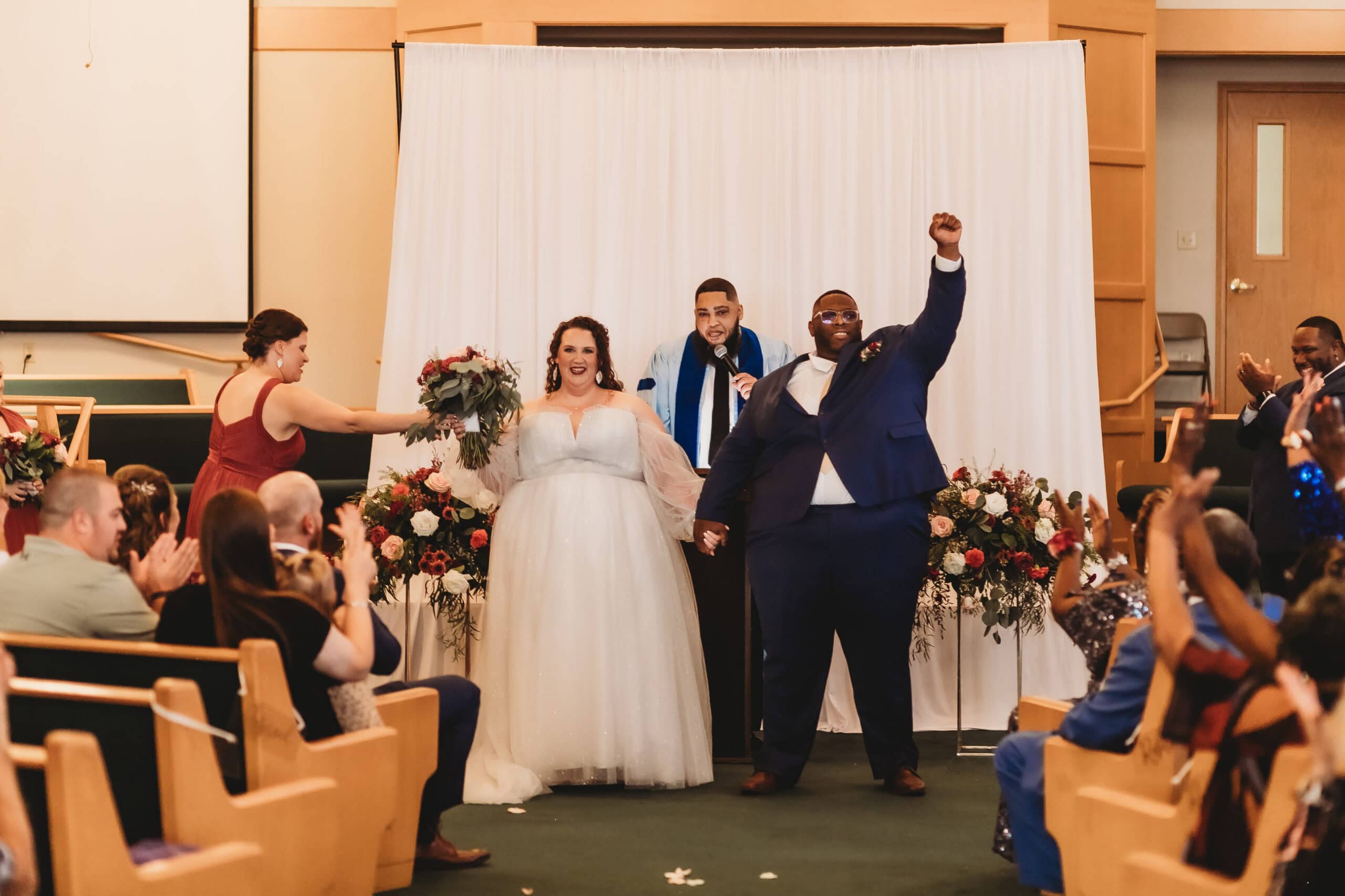 A joyful bride in a sparkling plus size bridal gown and her groom celebrate their wedding ceremony with raised hands, surrounded by cheers and floral decor.
