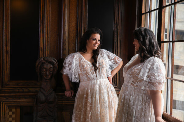 "Two plus-size brides wearing matching lace wedding gowns with flutter sleeves and intricate floral details, sharing a joyful moment in an elegant, wood-paneled venue with natural light streaming in."